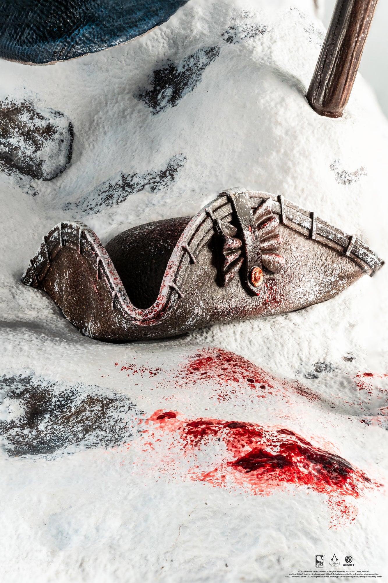 A detailed close-up of the snowy base of the Animus Connor statue, showing a discarded colonial hat and blood spatter on the snow.