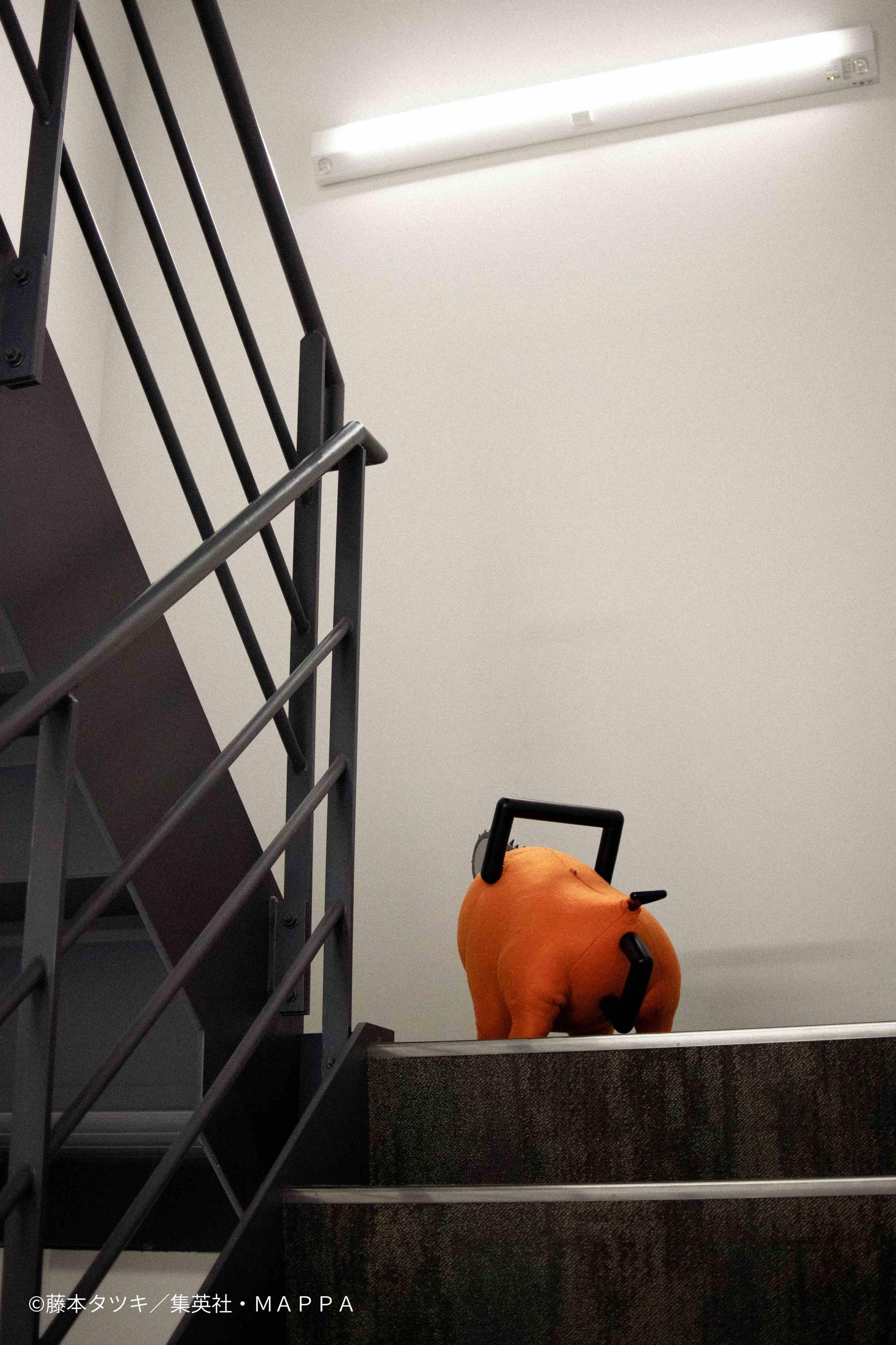 Rear view looking up at the Pochita life-size plush doll standing at the top of a staircase.