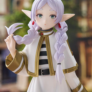 Close-up view of the Frieren figure displayed against a blurred fantasy village backdrop with European-style timber-framed houses.