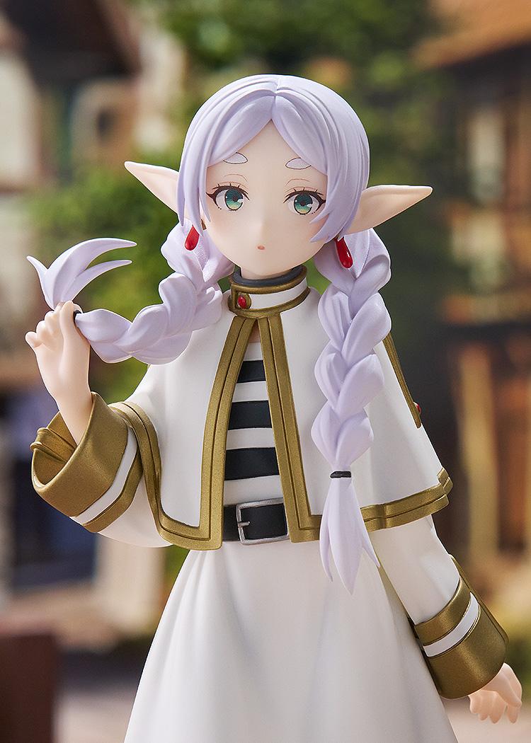 Close-up view of the Frieren figure displayed against a blurred fantasy village backdrop with European-style timber-framed houses.
