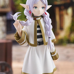 High-angle close-up portrait of the Good Smile Company Frieren figure with her signature neutral expression, set against an outdoor town background.