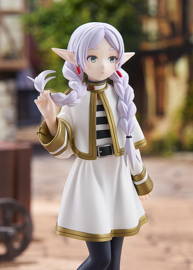 High-angle close-up portrait of the Good Smile Company Frieren figure with her signature neutral expression, set against an outdoor town background.