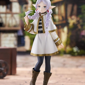 Full-body showcase of the Frieren Braids version figure standing on a stone-paved floor in a miniature medieval village setting.