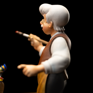 A side profile close-up of the Geppetto figure from the Iron Studios Pinocchio statue, showing him holding a paintbrush and pointing at the doll.