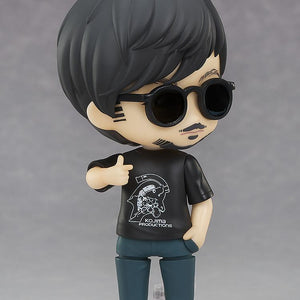 Three-quarter view of Hideo Kojima Nendoroid figure wearing sunglasses giving a thumbs up on display base.