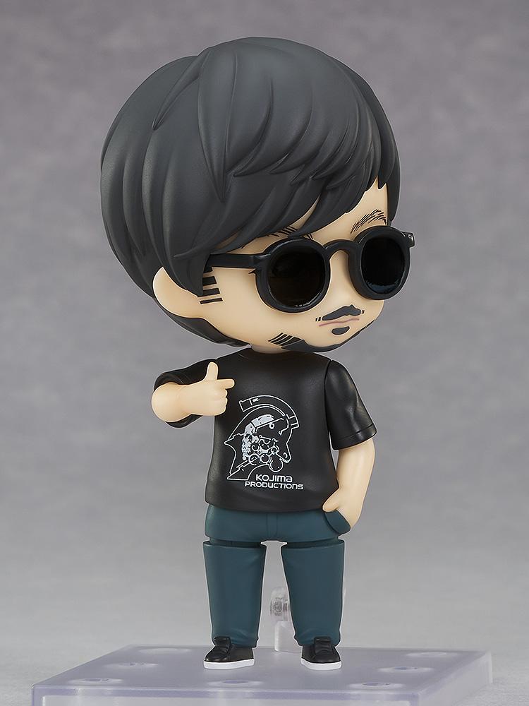 Three-quarter view of Hideo Kojima Nendoroid figure wearing sunglasses giving a thumbs up on display base.