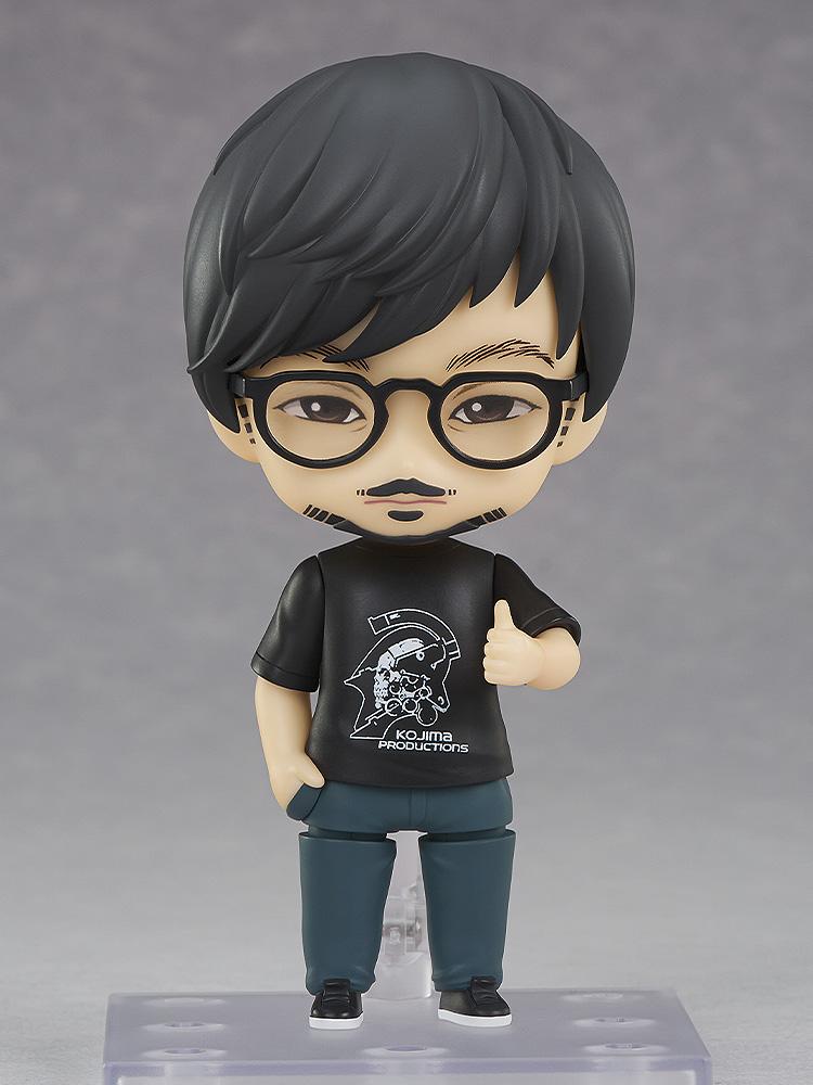 Good Smile Company Hideo Kojima Nendoroid figure wearing black Kojima Productions t-shirt and glasses in a thumbs-up pose on display stand.