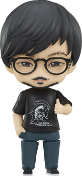 Studio cutout of Good Smile Company Hideo Kojima Nendoroid figure in logo t-shirt and glasses isolated on a white background.