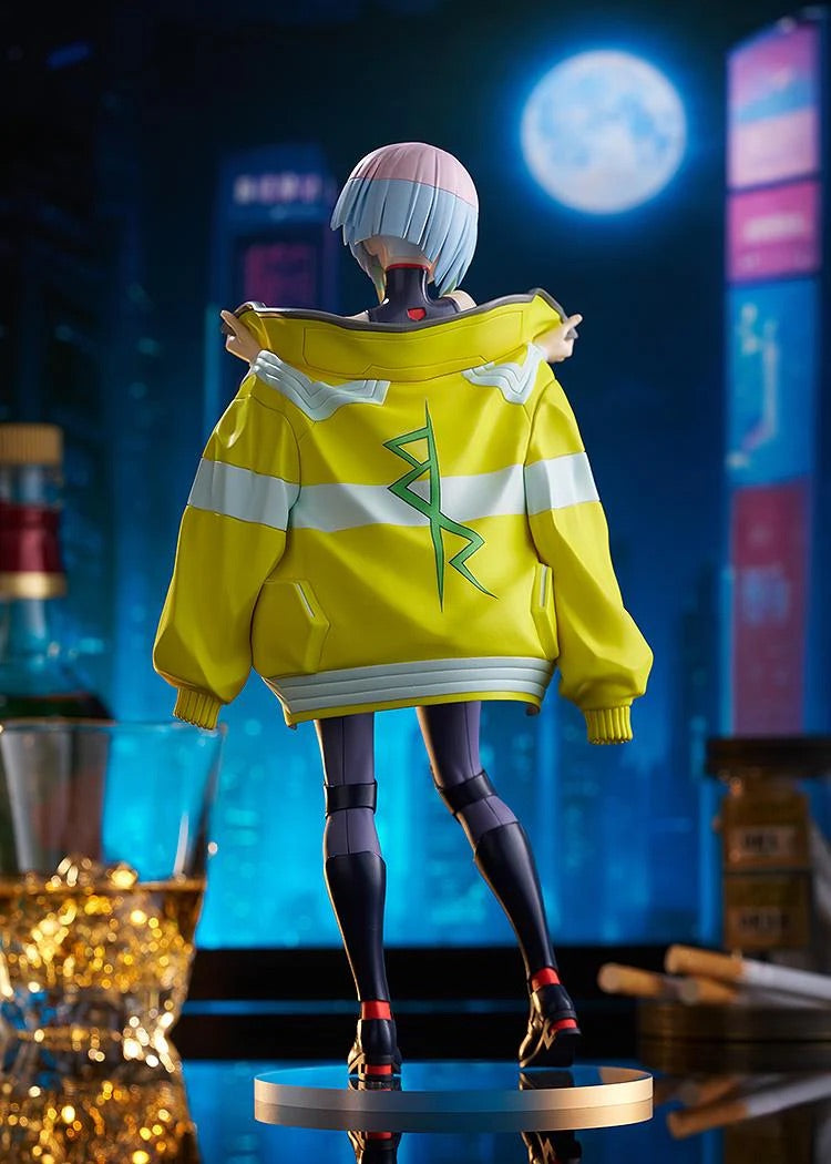 Rear view of the Lucy figure, showcasing the back of her oversized yellow jacket with the green symbol, set against a dark city skyline.
