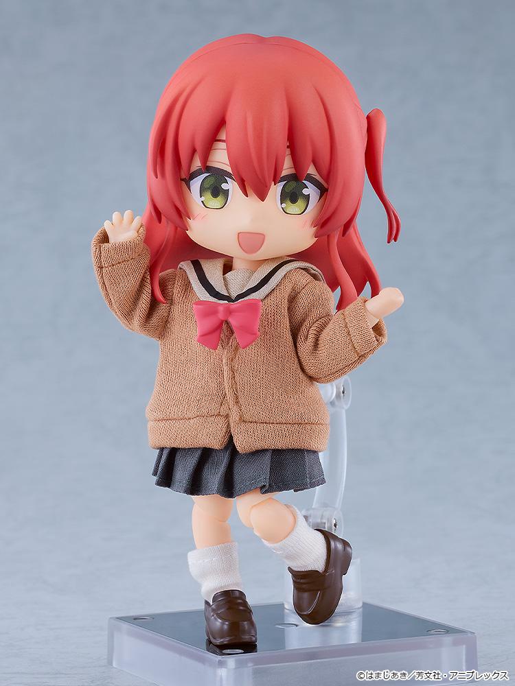 Full-body standing shot of the Ikuyo Kita Nendoroid Doll anime action figure waving cheerfully while mounted on a clear plastic display stand against a soft grey studio background.