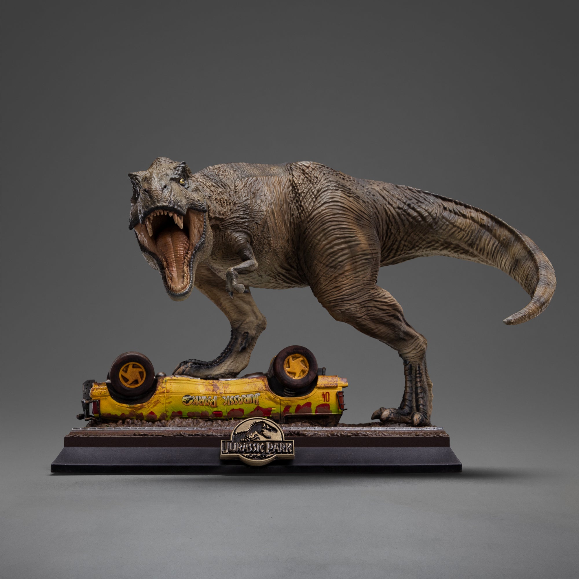 Front view of the Iron Studios Icons line T-Rex Attack polystone statue from Jurassic Park, featuring a Tyrannosaurus Rex roaring with an open mouth while stepping on an overturned yellow tour vehicle on a black base.