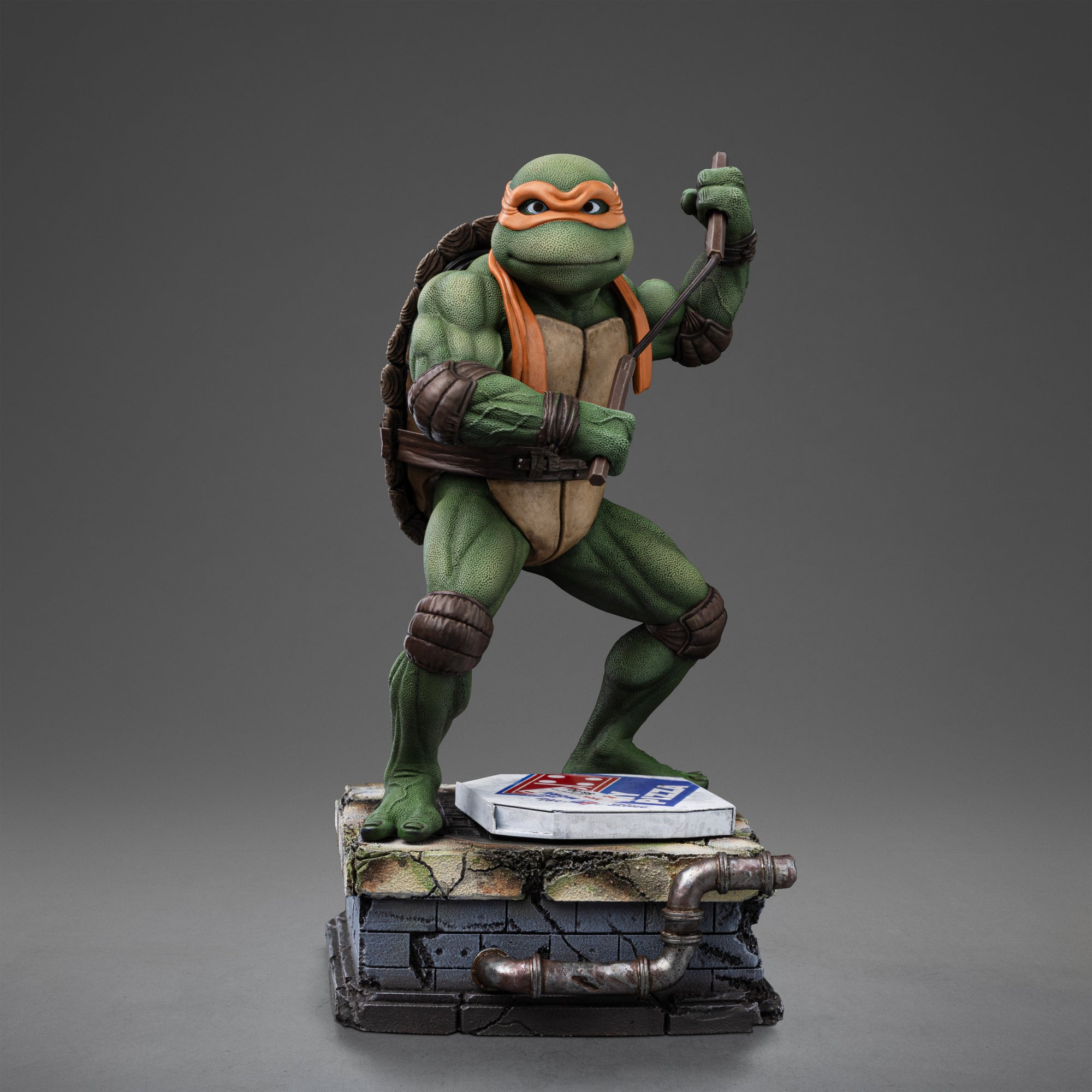 Full front view of the Iron Studios TMNT 1990 Michelangelo 1/10 scale statue poised with nunchaku above a sewer stone base with pizza box.