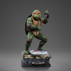 Full front view of the Iron Studios TMNT 1990 Michelangelo 1/10 scale statue poised with nunchaku above a sewer stone base with pizza box.