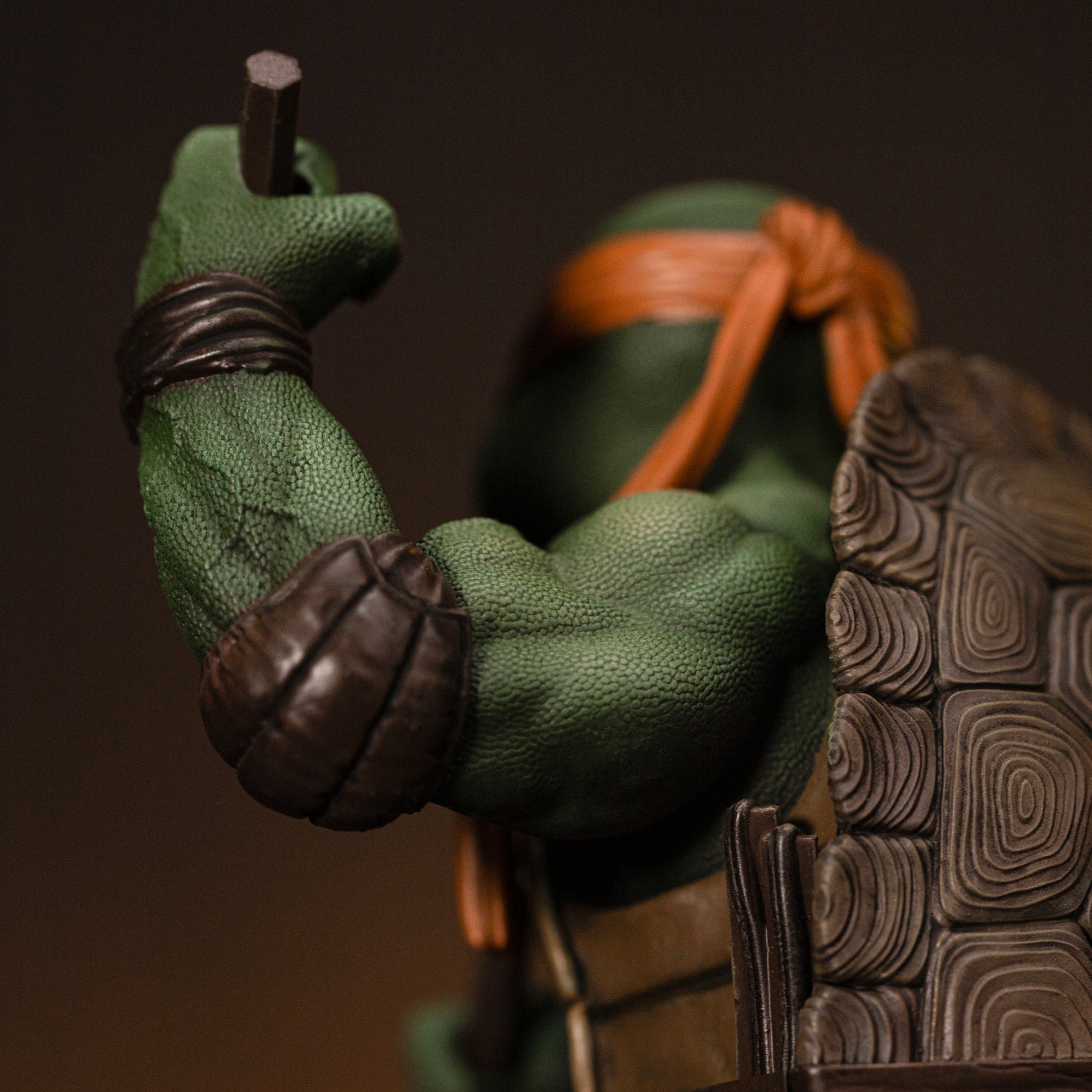 Rear three-quarter close-up focusing on Michelangelo's muscular shoulder, leather elbow pad, and nunchaku grip.