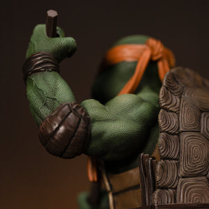Rear three-quarter close-up focusing on Michelangelo's muscular shoulder, leather elbow pad, and nunchaku grip.