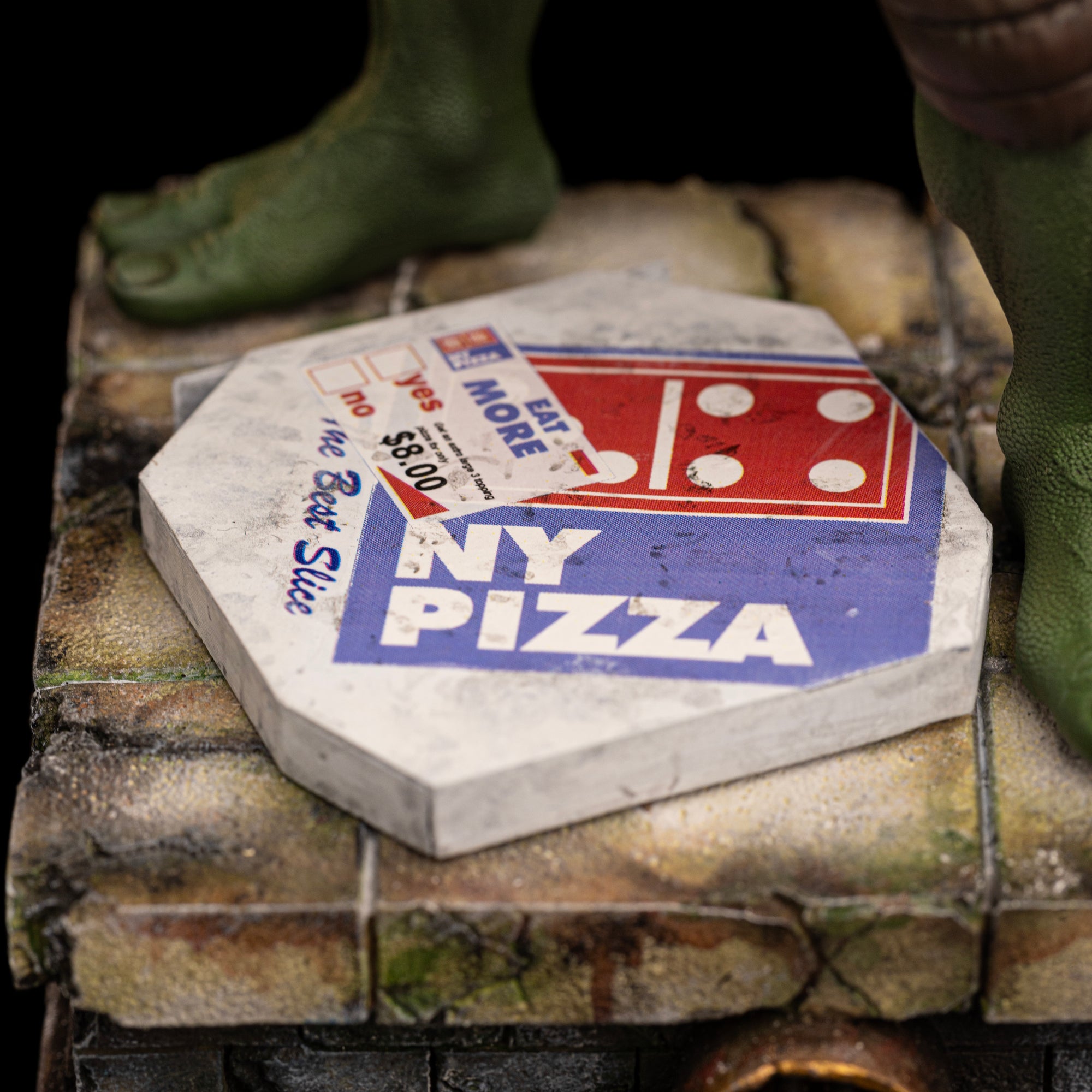 Close-up of the vintage NY Pizza box accessory resting on the weathered stone sewer base of the Iron Studios Raphael statue.