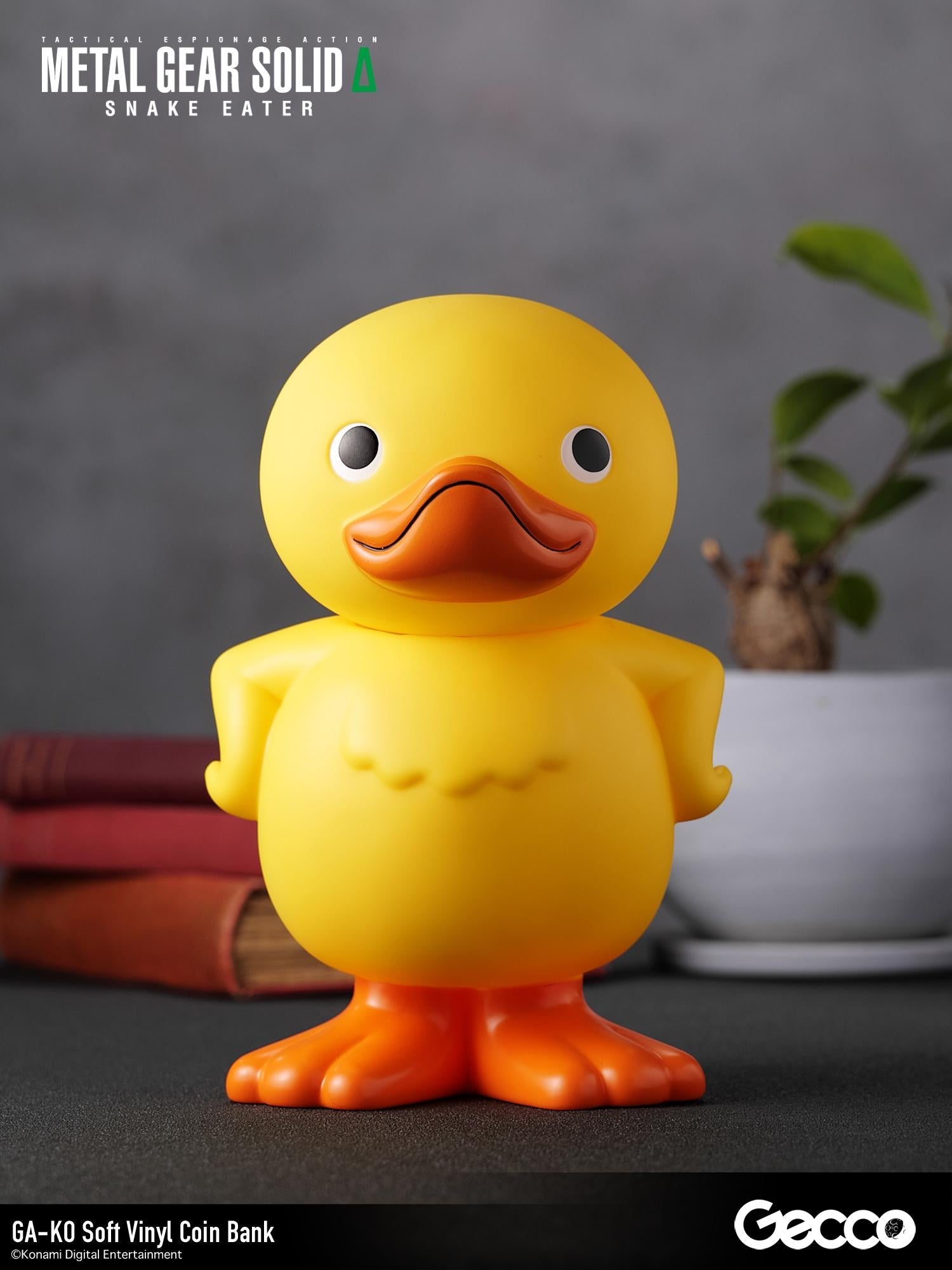 Front view of the GA-KO soft vinyl duck coin bank arranged on a desk display with books and houseplant.