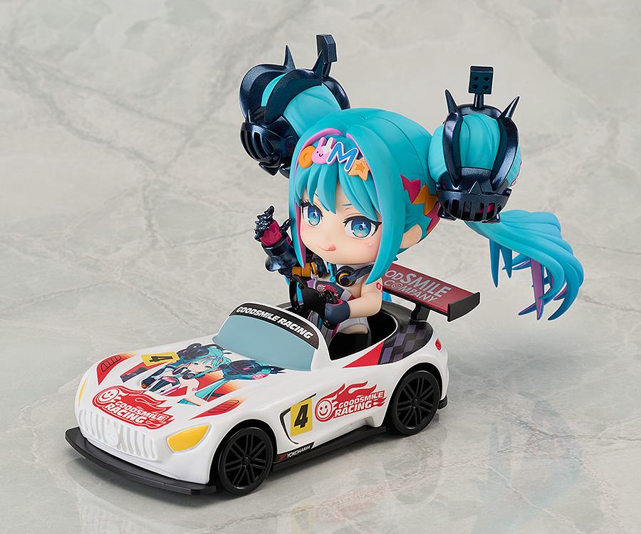 An action shot showing the Nendoroid Racing Miku: 2026 Ver. figure seated in and driving the included Nendoroid car on a racetrack, smiling and waving.