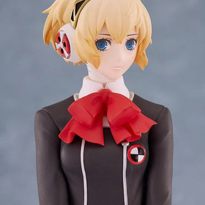 Close-up on the face and upper chest of the Aigis figure against a muted background, focusing on her expression and the red bow.