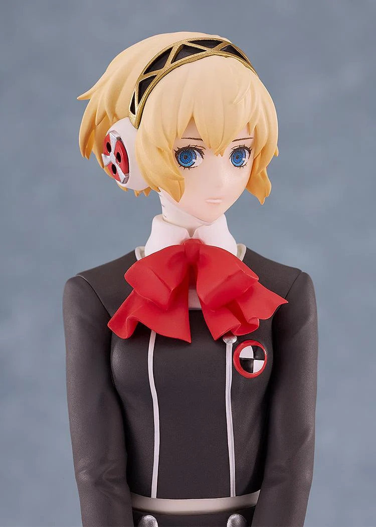 Close-up on the face and upper chest of the Aigis figure against a muted background, focusing on her expression and the red bow.