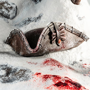 A detailed close-up of the snowy base of the Animus Connor statue, showing a discarded colonial hat and blood spatter on the snow.