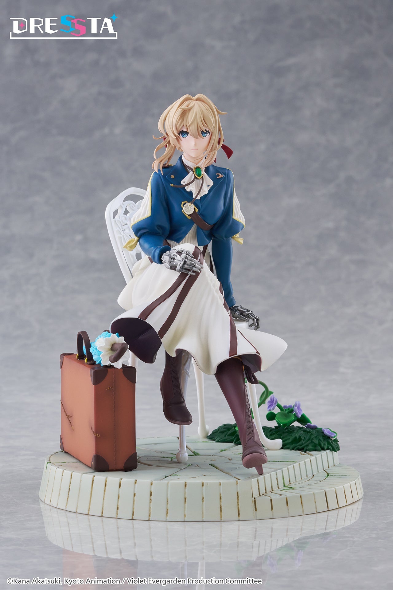 Full front view of Taito Violet Evergarden DRESSTA figure sitting on a white chair next to a suitcase on a stone base